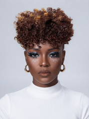 A close-up portrait of a model wearing a short, voluminous curly wig with dark brown and warm caramel-blonde highlights, styled with dramatic eye makeup and gold hoop earrings against a light gray background.