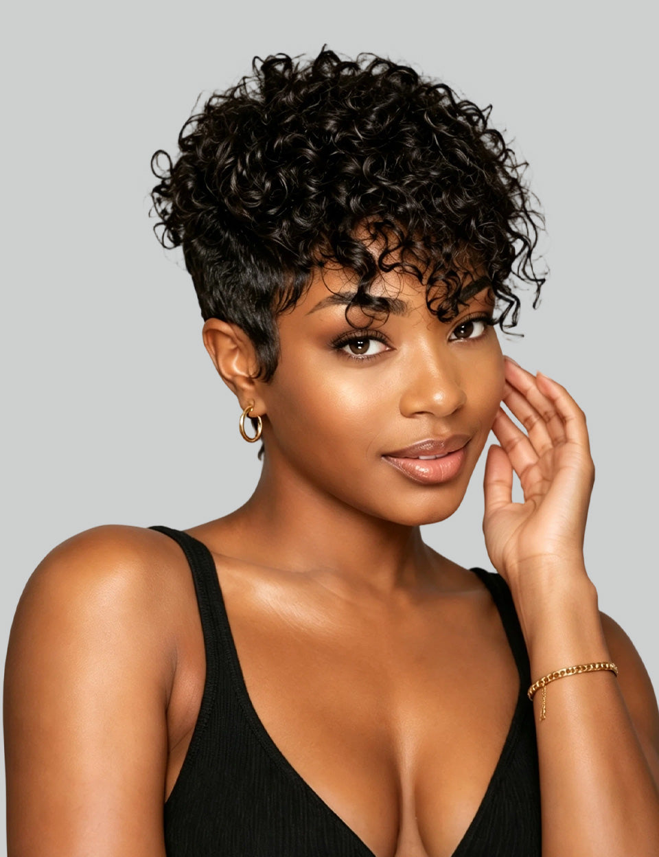 A woman with short, curly black hair and a side-swept undercut hairstyle, wearing a black ribbed tank top against a solid gray background.