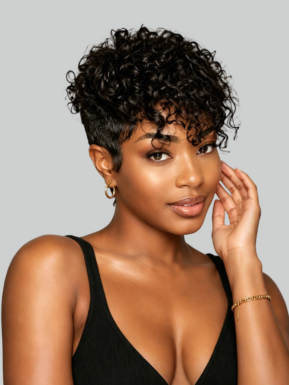 A woman with short, curly black hair and a side-swept undercut hairstyle, wearing a black ribbed tank top against a solid gray background.