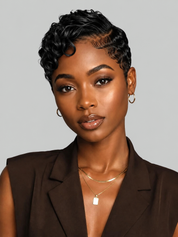 A close-up portrait of a woman with short, curly black hair wearing a dark brown sleeveless blazer, layered gold necklaces, and hoop earrings against a solid light gray background.