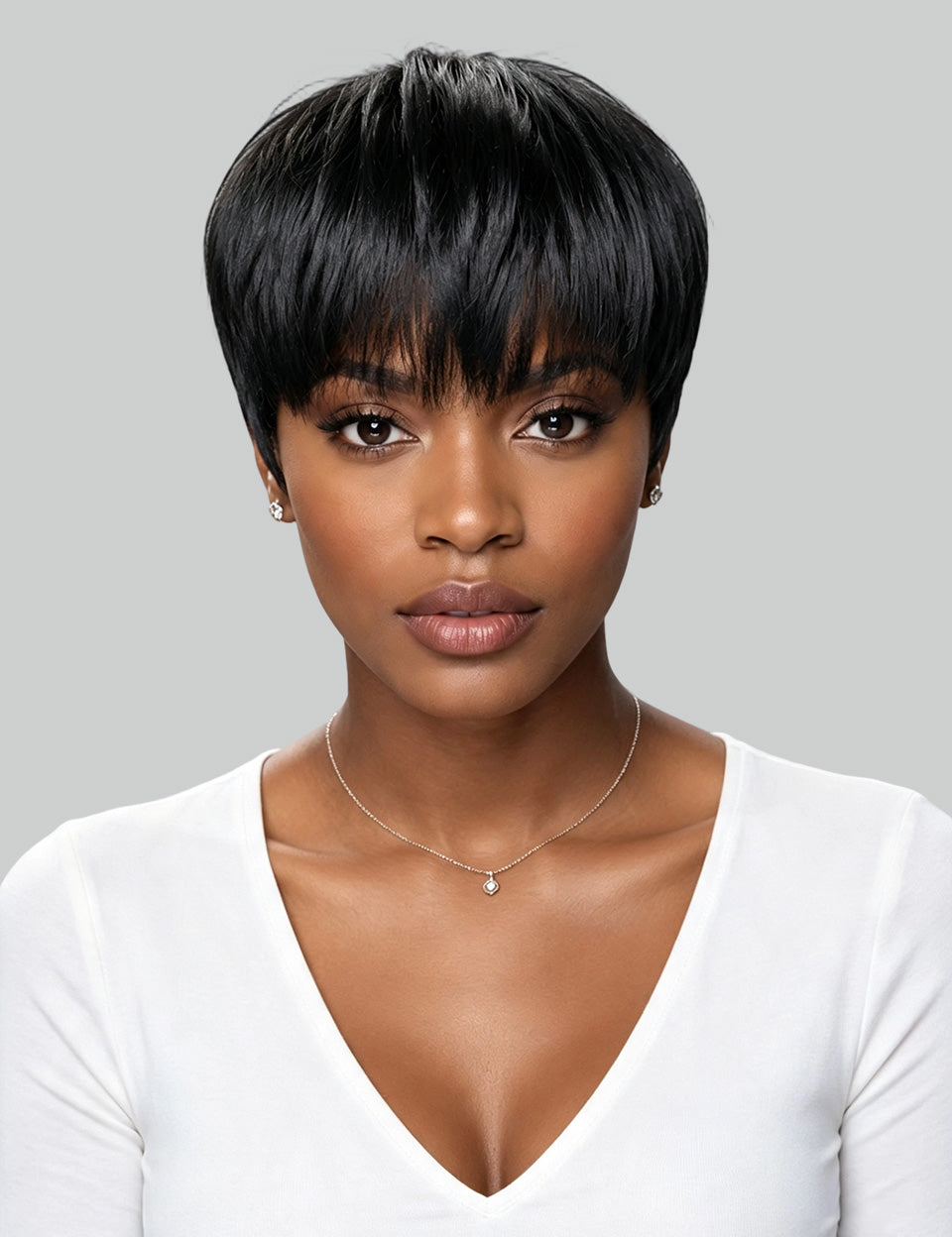 A woman with a short, straight black wig or hairpiece shown from the chest up against a light gray studio background. She is wearing a white V-neck top and a delicate necklace.