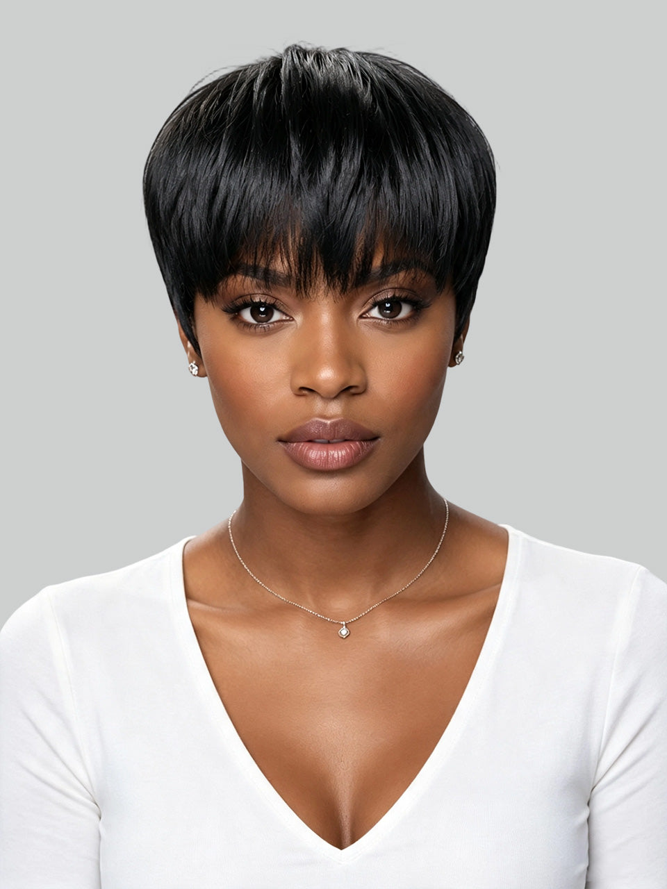 A woman with a short, straight black wig or hairpiece shown from the chest up against a light gray studio background. She is wearing a white V-neck top and a delicate necklace.