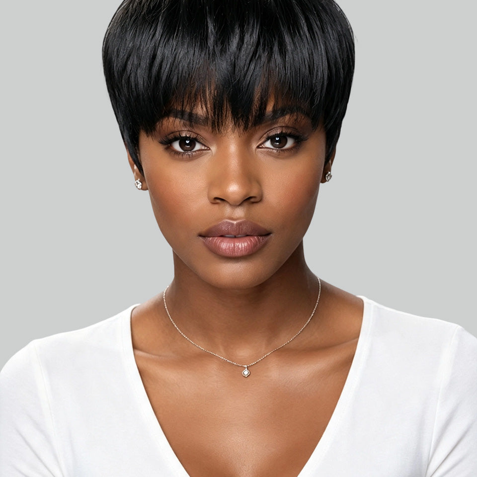 A woman with a short, straight black wig or hairpiece shown from the chest up against a light gray studio background. She is wearing a white V-neck top and a delicate necklace.