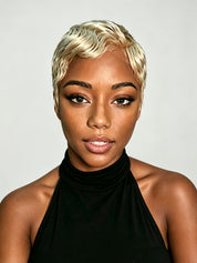 A close-up portrait of a woman with a short, textured platinum blonde wig, wearing a black halter top against a light gray background.