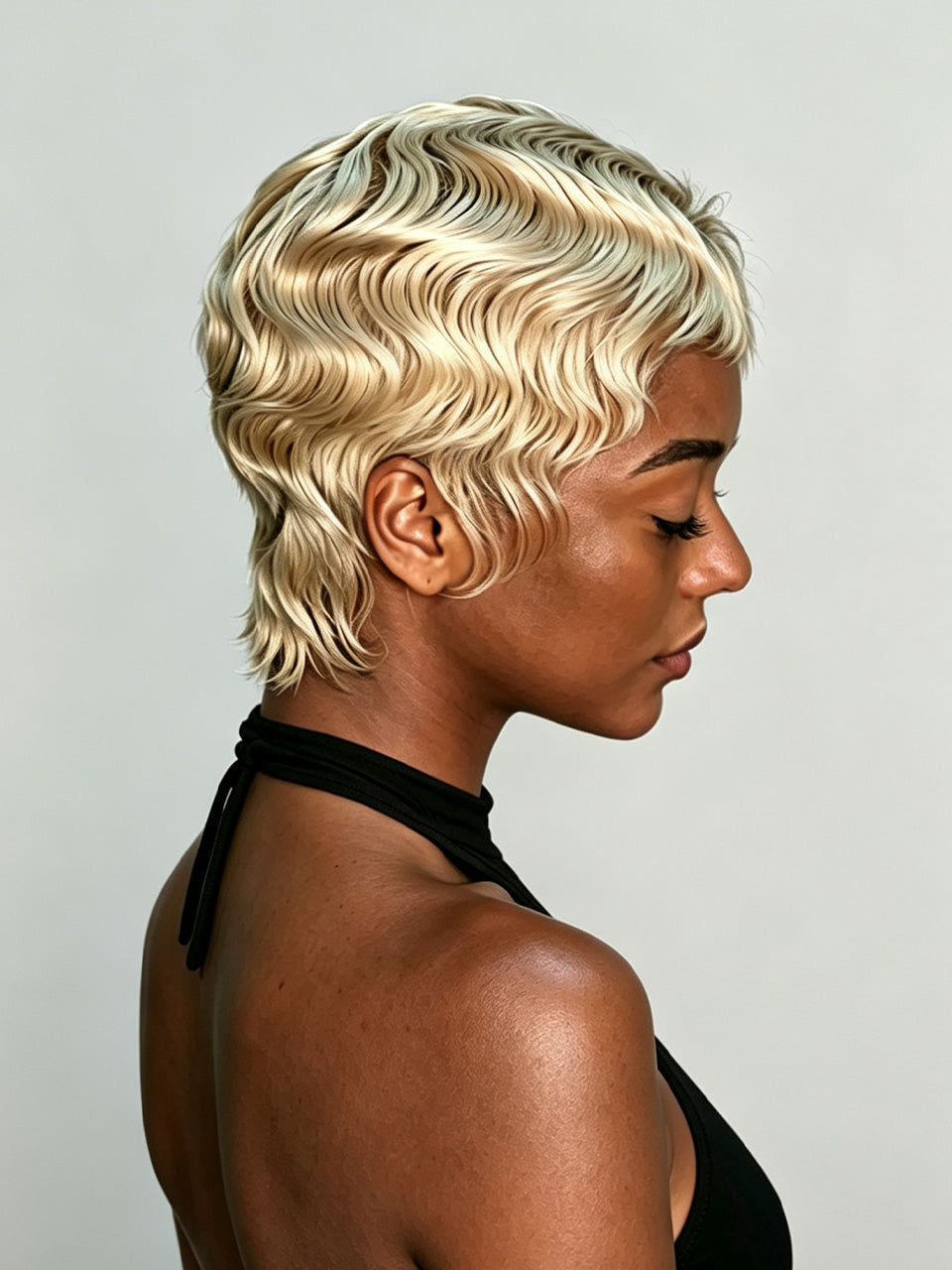 A woman shown in side profile wearing a short, wavy platinum blonde wig against a plain light gray background.