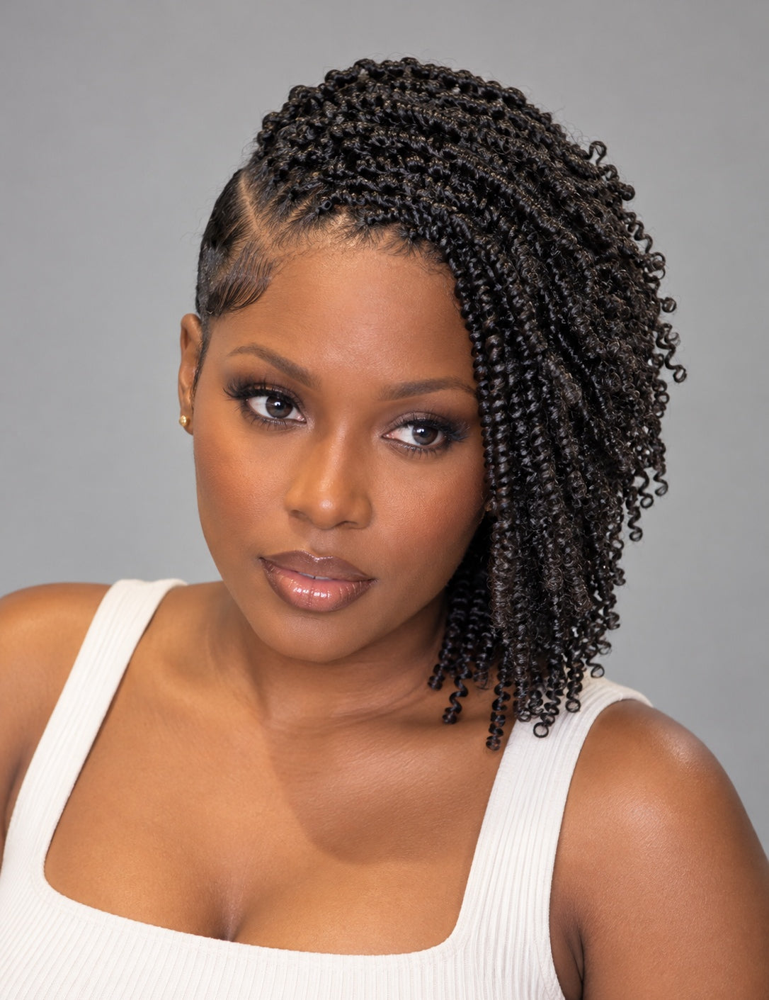 A woman with dark brown to black curly hair styled in a side braid, wearing a white ribbed tank top against a solid gray studio background.