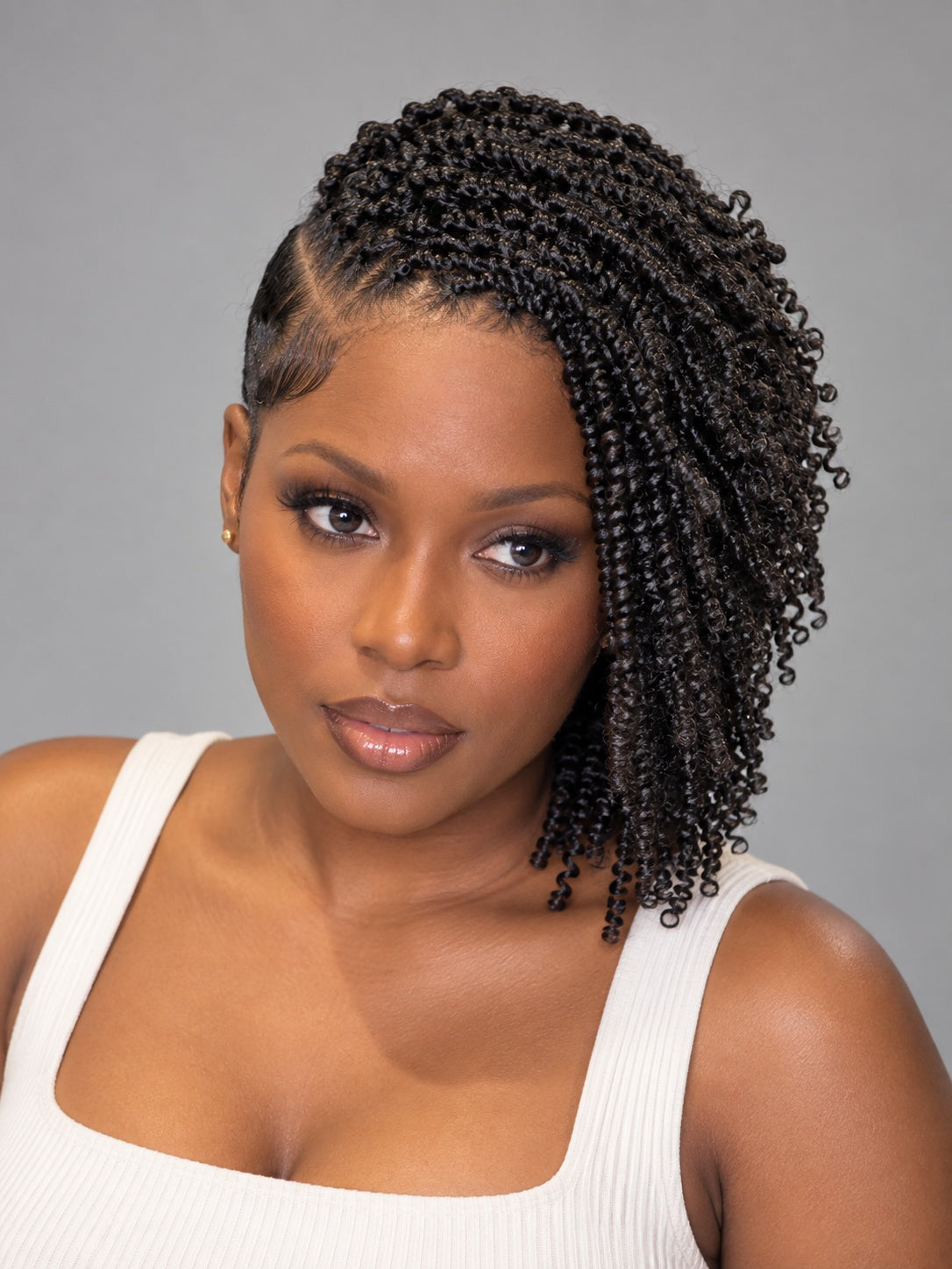 A woman with dark brown to black curly hair styled in a side braid, wearing a white ribbed tank top against a solid gray studio background.