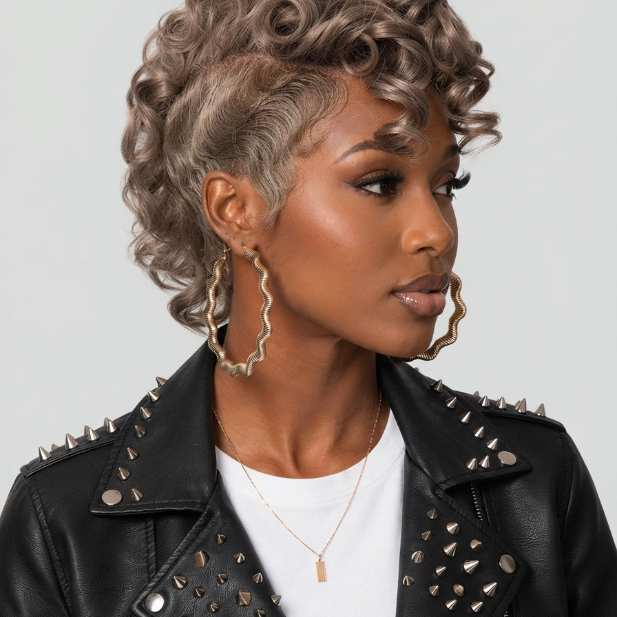 A woman wearing a short, voluminous curly wig with a cool ash-blonde or silver-gray tone, styled with a side part. She is wearing a black studded leather jacket, a white t-shirt, and large gold hoop earrings against a solid light gray background.