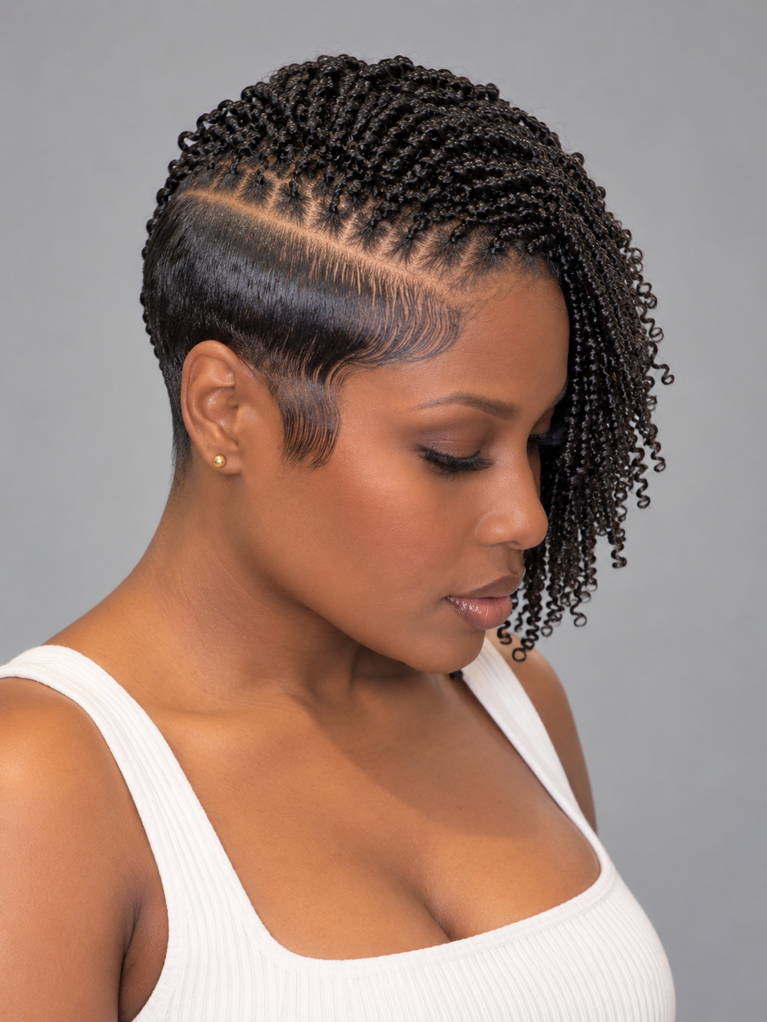 A woman with a short, styled hairstyle featuring dark curly cornrow braids and a clean side part, wearing a white ribbed tank top against a solid gray background.