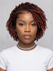 A woman with short, tight, reddish-brown box braids or twists wearing a white t-shirt and a sparkly silver choker, set against a neutral gray background.