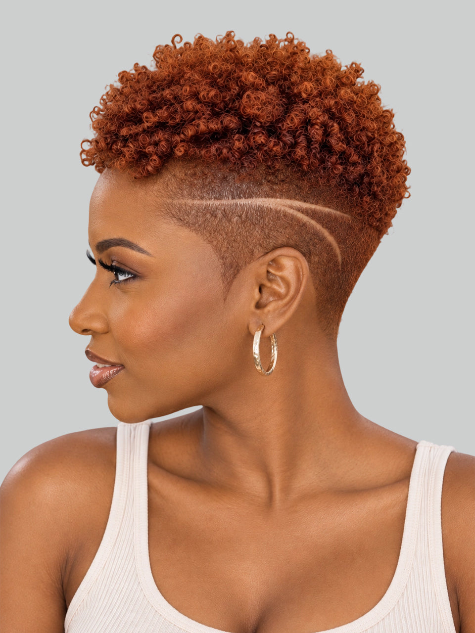 A woman in profile wearing a short, curly auburn wig with a high undercut and geometric line detail on the side, paired with gold hoop earrings and a white ribbed tank top, against a solid gray background.