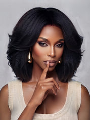 Frontal close-up of a woman wearing a black shoulder-length wig with soft, bouncy waves and a center part. The voluminous curls frame the face elegantly, complemented by bold smoky eye makeup and gold teardrop earrings for a glamorous, confident look.