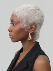 A woman in profile wearing a short, textured platinum blonde wig, black cat-eye sunglasses, gold teardrop earrings, and a black sleeveless top against a solid gray background.
