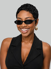 A smiling woman with short dark curly hair wears black cat-eye sunglasses and a black sleeveless blazer against a solid light gray background.