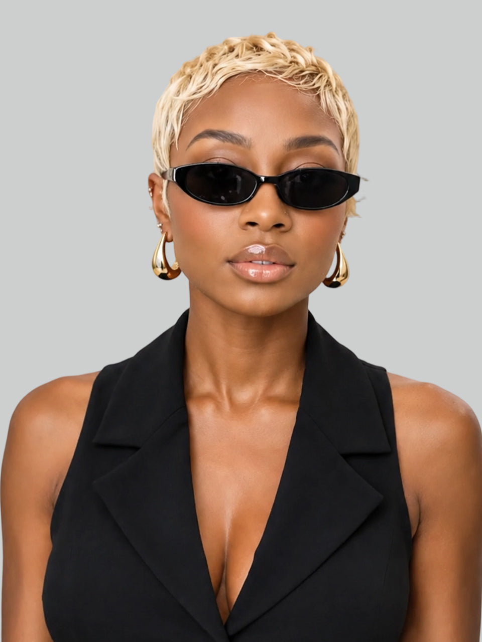A model with short blonde braided hair wearing black cat-eye sunglasses, gold hoop earrings, and a black sleeveless blazer against a solid gray background.