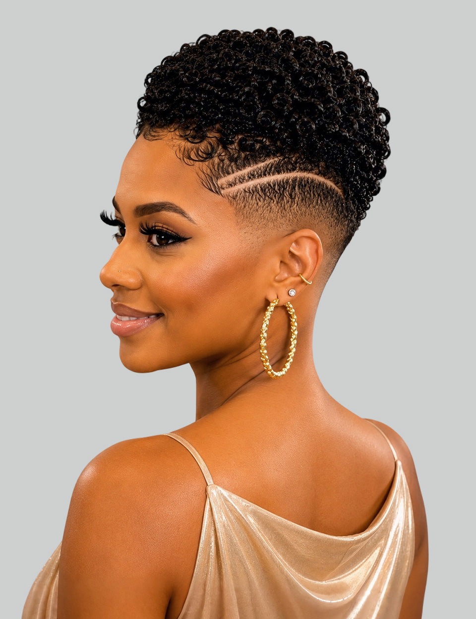 A woman shown in profile wearing a short, curly black wig with a high fade and geometric side haircut, paired with large gold hoop earrings and a champagne satin top against a light gray background.