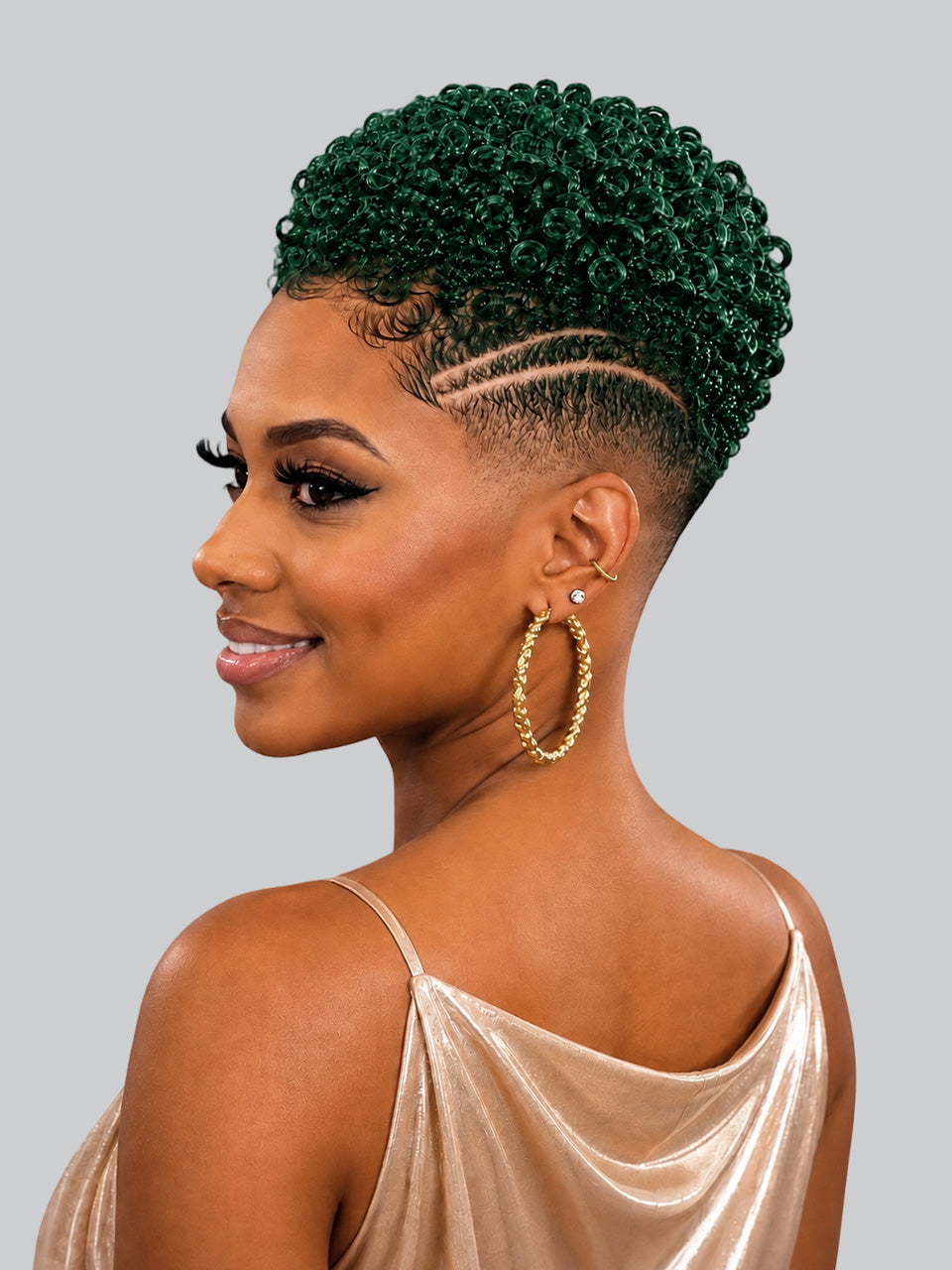 A woman shown in profile with a short, tightly curled hairstyle in deep emerald green and a faded undercut. She is wearing large gold hoop earrings and a champagne satin top against a plain light gray studio background.