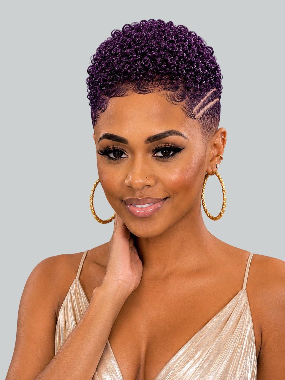 A woman with a short, tightly curled purple wig and gold hoop earrings, wearing a champagne-colored pleated top against a solid gray background.