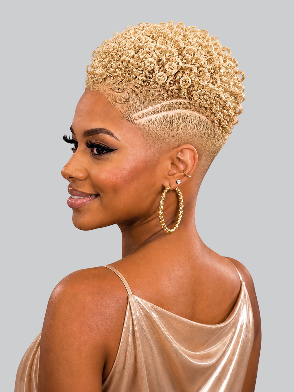 A woman shown in side profile wearing a short, tightly curled blonde wig with a faded undercut and geometric side detailing. She is wearing a champagne satin top and large gold hoop earrings against a solid gray studio background.