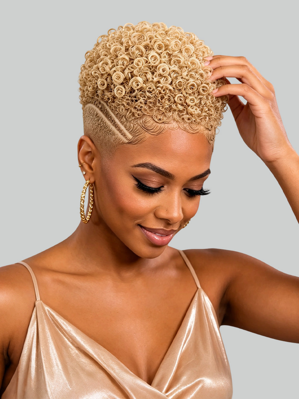A woman with a short, tightly curled blonde wig or hairpiece shown from the shoulders up against a plain light gray studio background. She is wearing a shiny champagne satin top and large gold hoop earrings, and her hand is touching the hair.