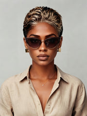 Frontal portrait of a model wearing a black-and-blonde pixie wig with wet-look texture, paired with cat-eye sunglasses and gold square earrings. The contrast between dark roots and blonde highlights adds dimension, while the neutral beige shirt keeps the focus on the bold hairstyle and accessories.