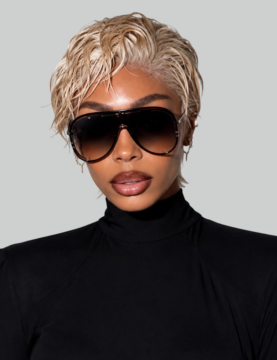 Frontal portrait of a model in a black turtleneck wearing a short golden blonde human hair wig. The style features a textured, wet-and-wavy slicked-back finish paired with oversized sunglasses.
