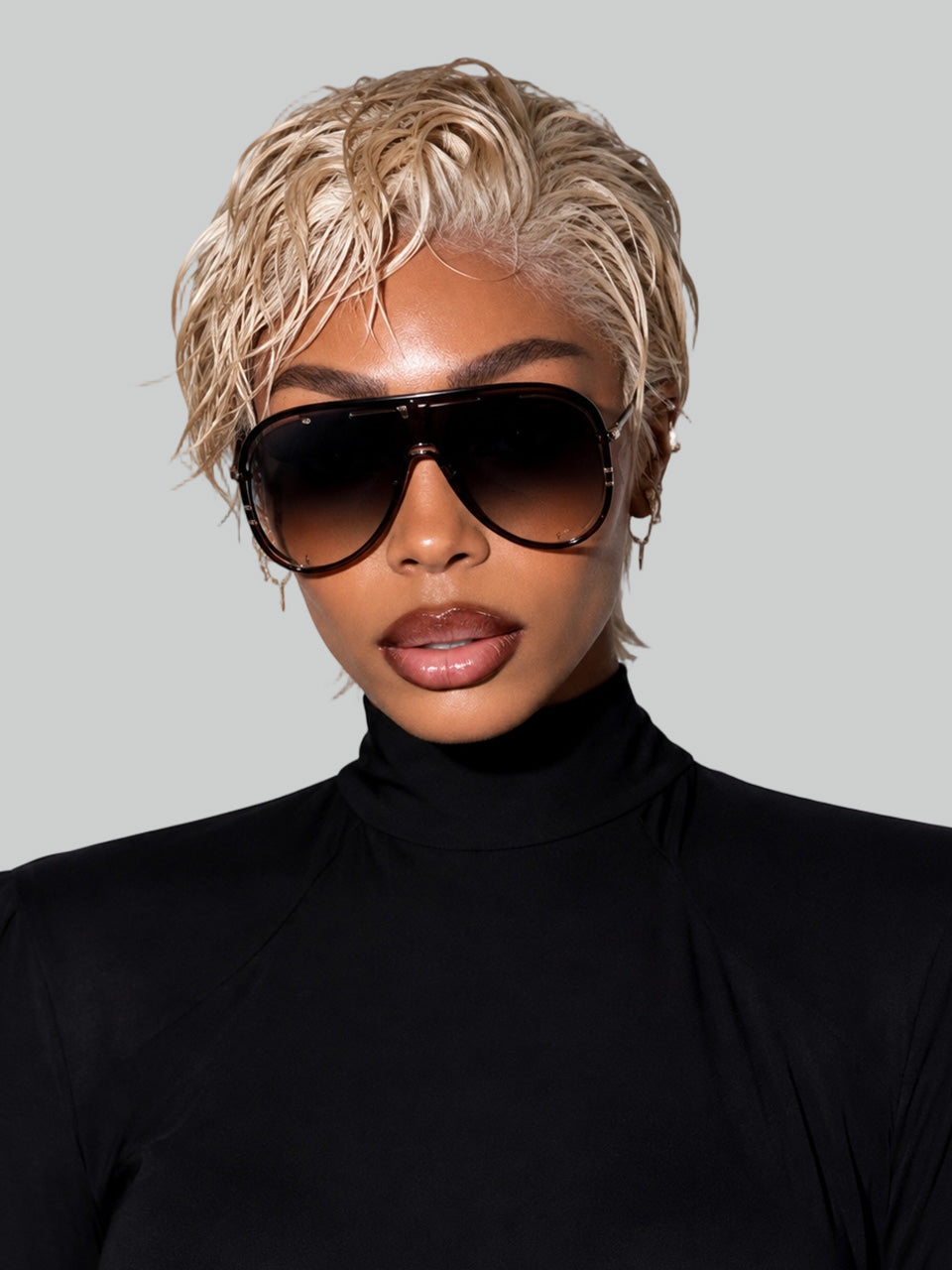 Frontal portrait of a model in a black turtleneck wearing a short golden blonde human hair wig. The style features a textured, wet-and-wavy slicked-back finish paired with oversized sunglasses.