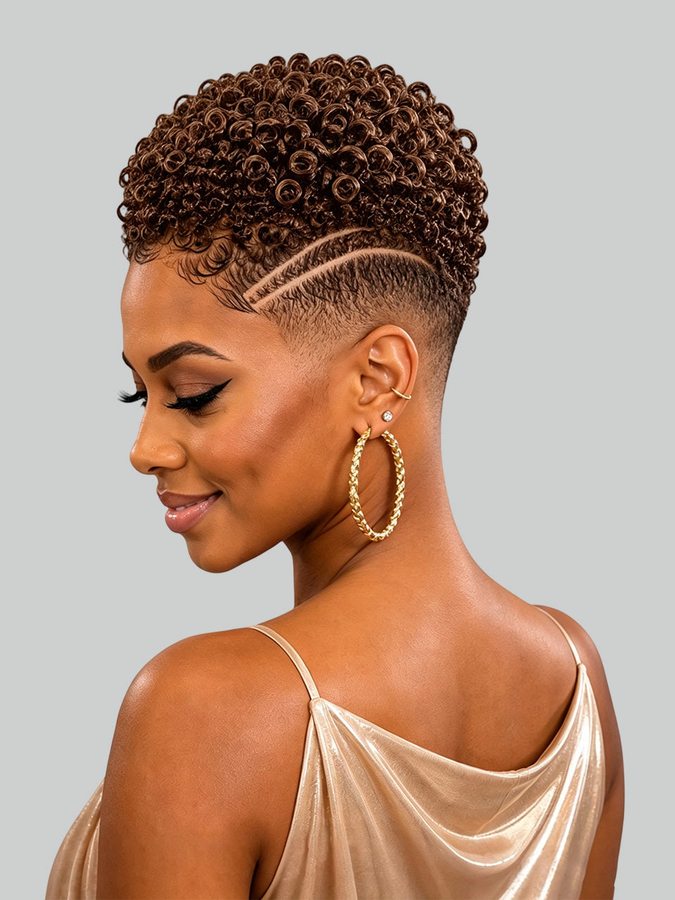 A woman shown in profile with short, tightly curled brown hair and a faded undercut. She is wearing large gold hoop earrings and a champagne satin top against a plain light gray studio background.