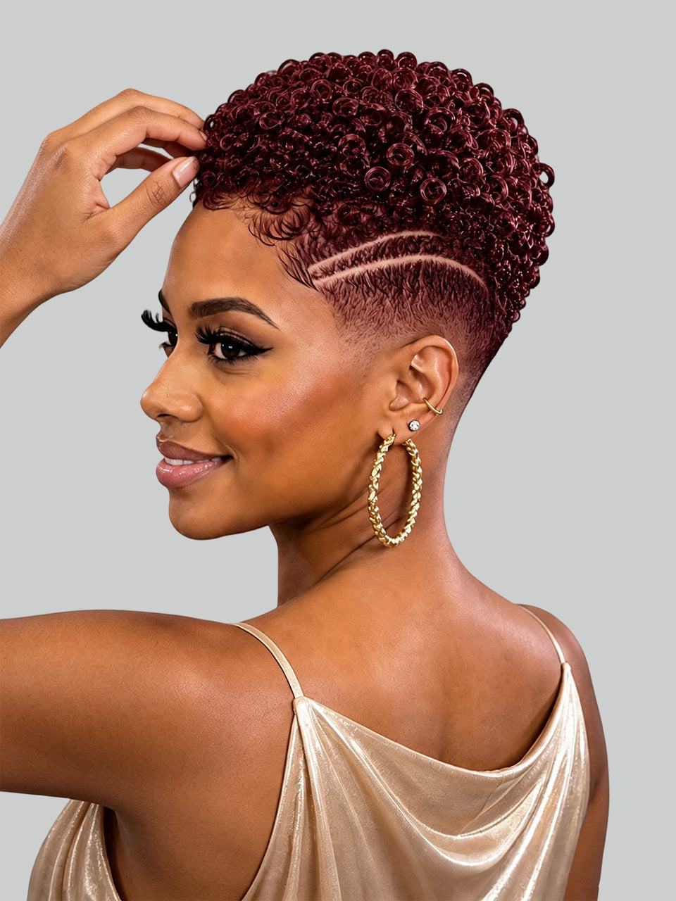 A woman with a short, curly, deep burgundy wig is shown in profile against a solid gray background. She is wearing a champagne-colored satin top and large gold hoop earrings.