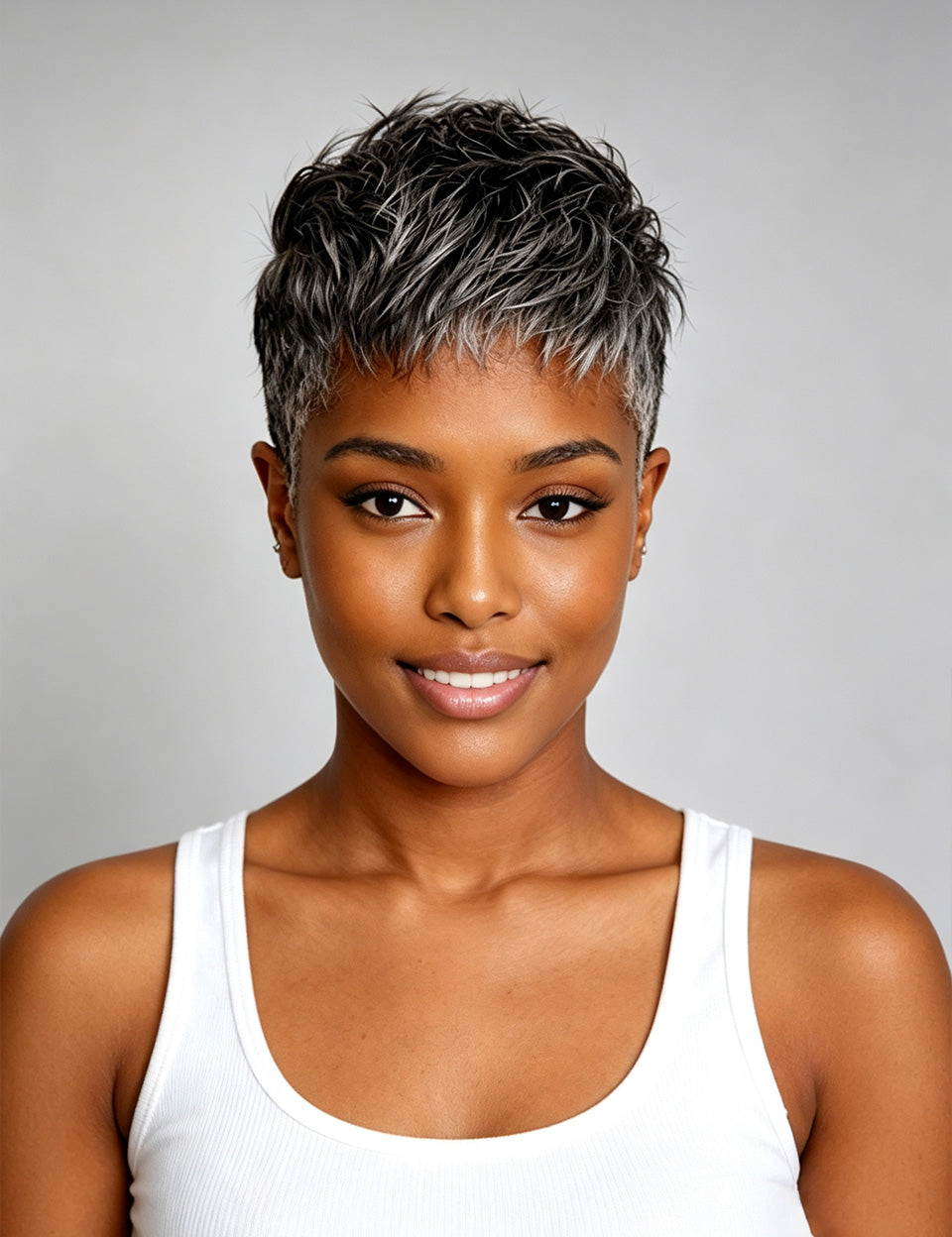 A woman with a short, textured pixie cut featuring dark brown and silver-gray highlights, wearing a white ribbed tank top against a solid gray studio background.