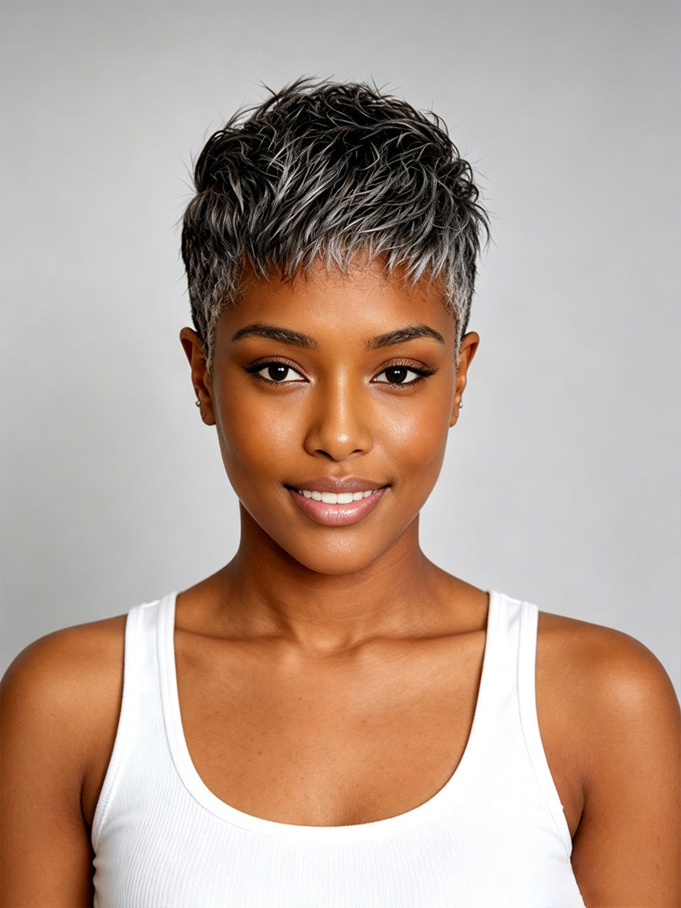 A woman with a short, textured pixie cut featuring dark brown and silver-gray highlights, wearing a white ribbed tank top against a solid gray studio background.