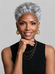 Frontal angled view of a woman wearing a silver grey short curly pixie wig with tight spiral curls. The model rests her chin on her hand, highlighting the wig’s voluminous crown and natural hairline, perfect for a bold and confident look.