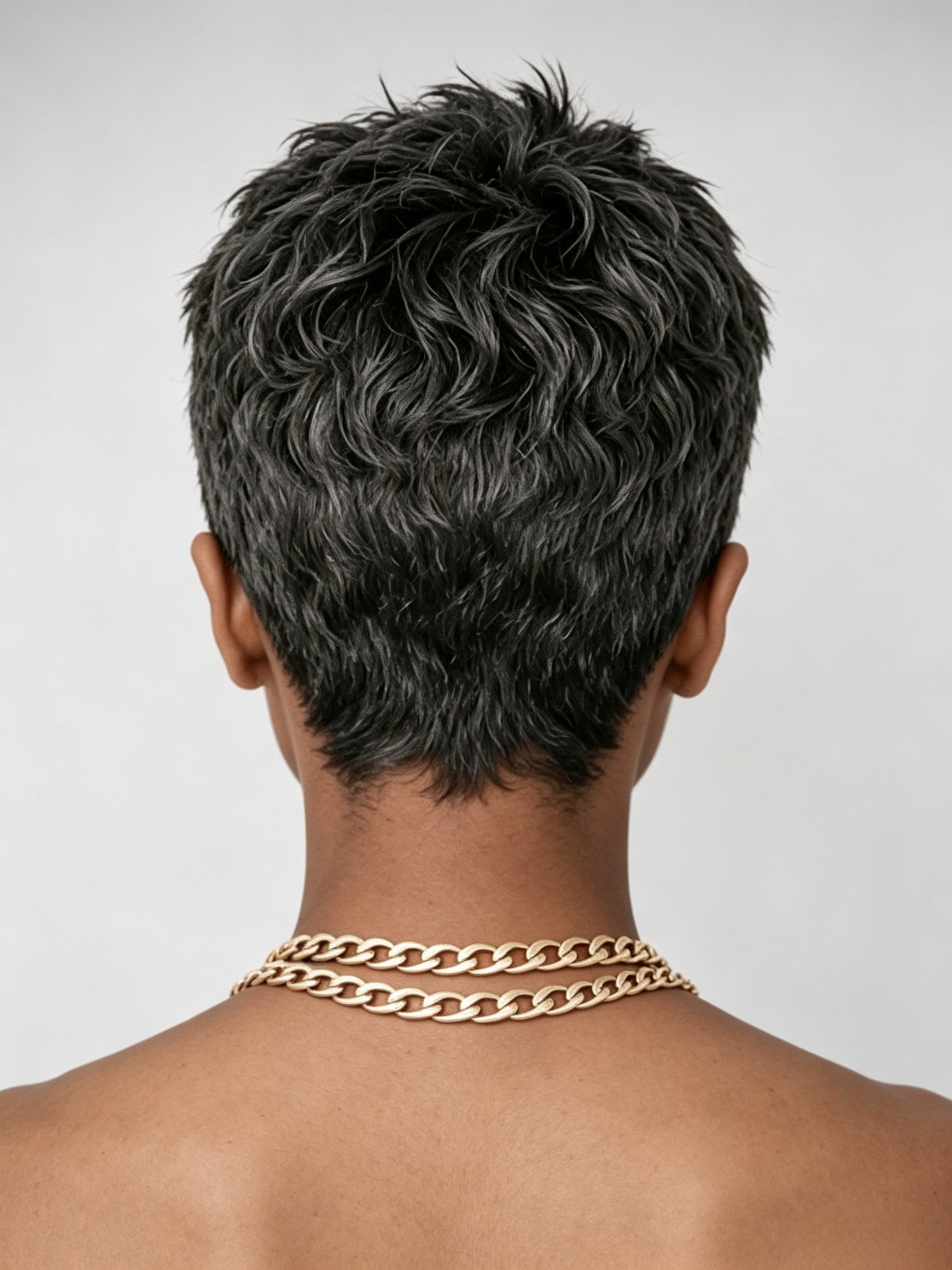 Back view of a woman wearing a short curly pixie wig with salt and pepper highlights. The image reveals the natural wave pattern and tapered nape, demonstrating how the wig sits neatly at the neckline with a gold chain choker accent.