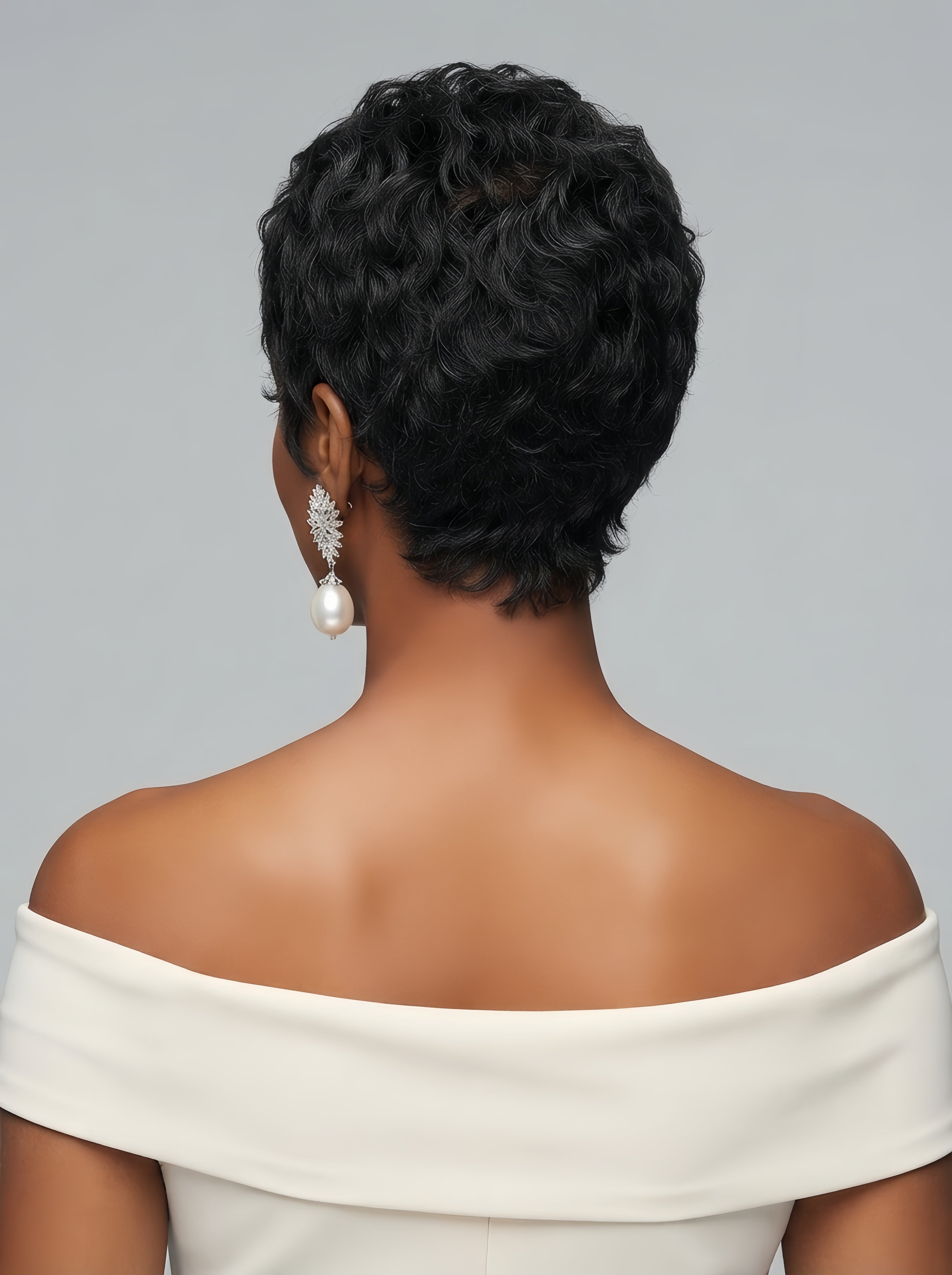 Back view of a woman wearing a short curly pixie wig in black with subtle grey highlights. The image highlights the tapered nape and the dense curl pattern at the crown, complemented by elegant pearl drop earrings and an off-shoulder white dress.