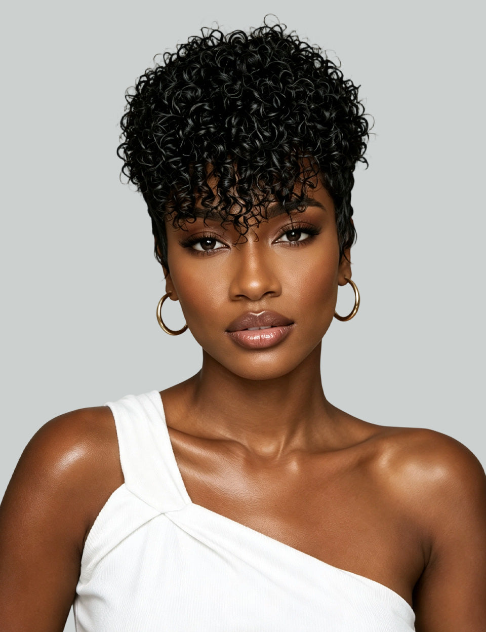 A woman with short, curly black hair wearing a white one-shoulder top and gold hoop earrings, photographed against a light gray studio background.
