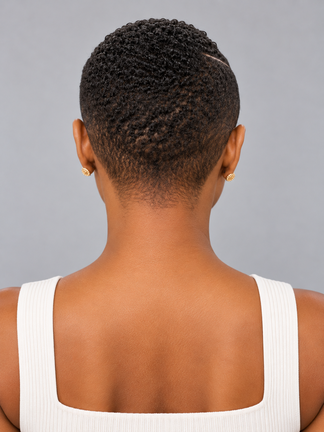 Back view of a person with short, textured black hair wearing a white ribbed tank top and small gold stud earrings against a plain gray background.