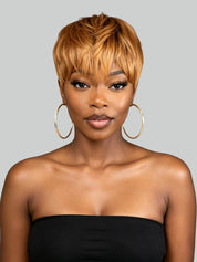 A woman with a short, layered, textured wig in warm caramel and golden blonde tones, styled with side-swept bangs. She is wearing large gold hoop earrings and a black strapless top against a solid light gray background.