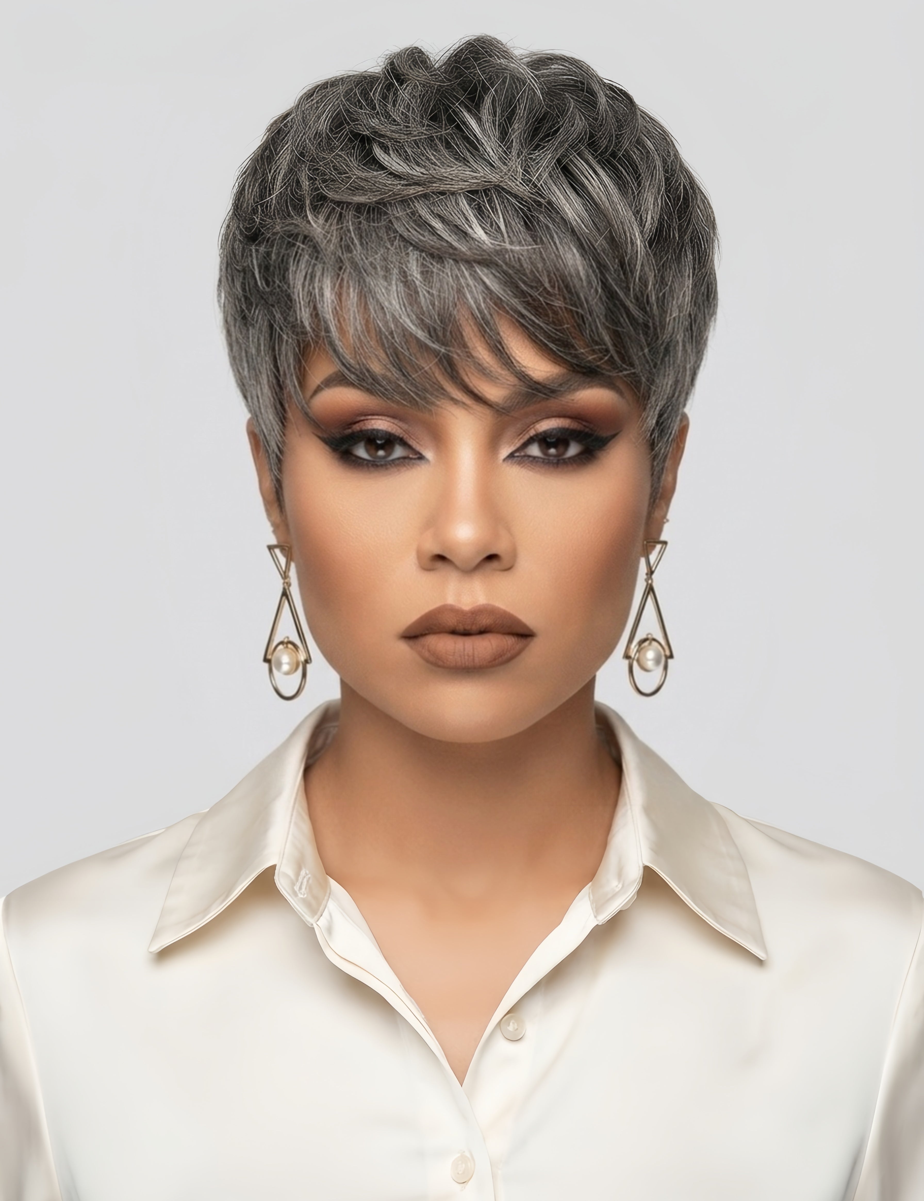Frontal close-up of a woman wearing a salt and pepper short hair topper with textured, layered bangs. The natural grey highlights blend seamlessly with dark roots, creating a realistic look. She pairs the style with warm brown eyeshadow, nude lipstick, and geometric pearl drop earrings for a sophisticated, professional appearance.