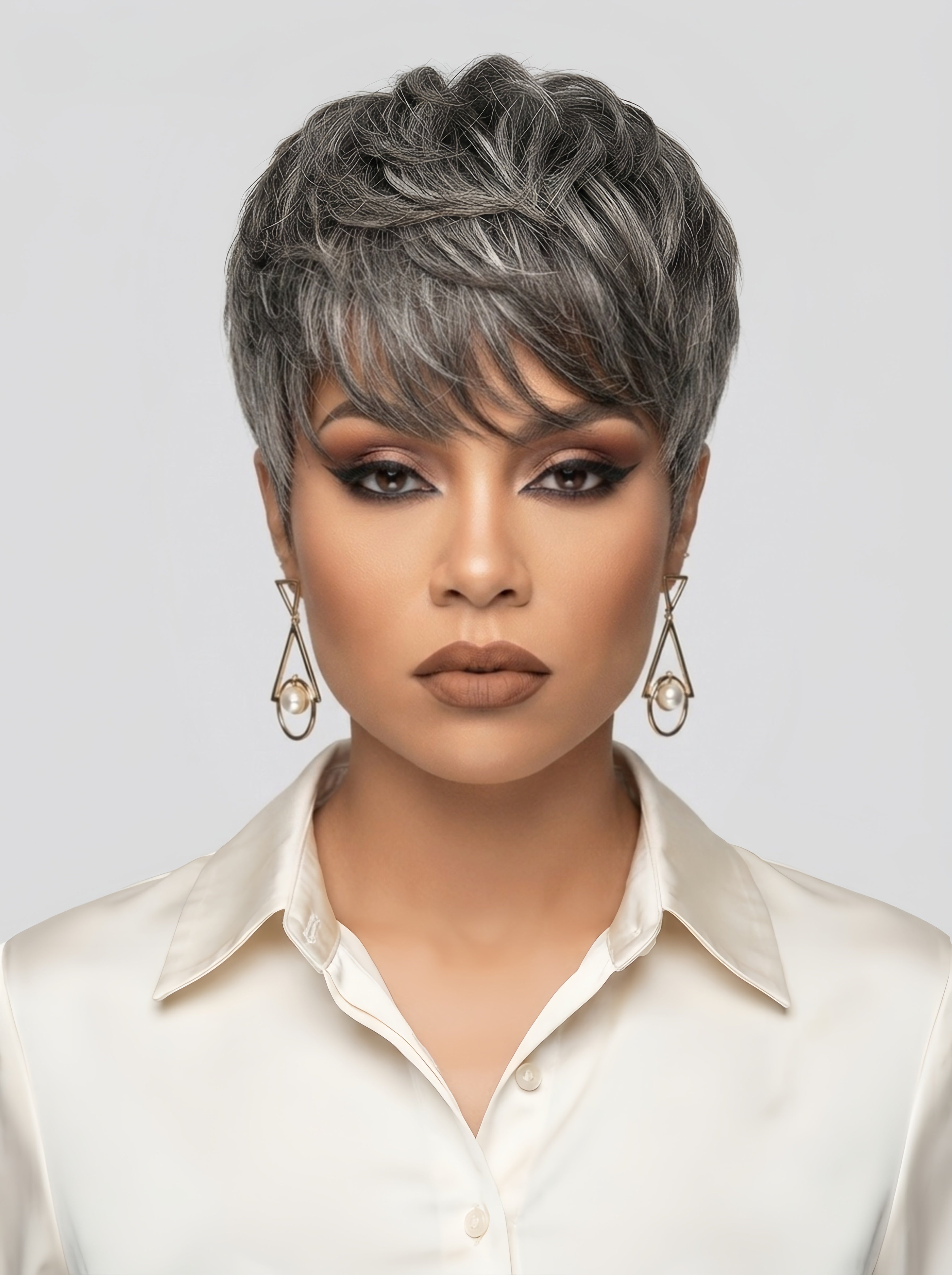 Frontal close-up of a woman wearing a salt and pepper short hair topper with textured, layered bangs. The natural grey highlights blend seamlessly with dark roots, creating a realistic look. She pairs the style with warm brown eyeshadow, nude lipstick, and geometric pearl drop earrings for a sophisticated, professional appearance.