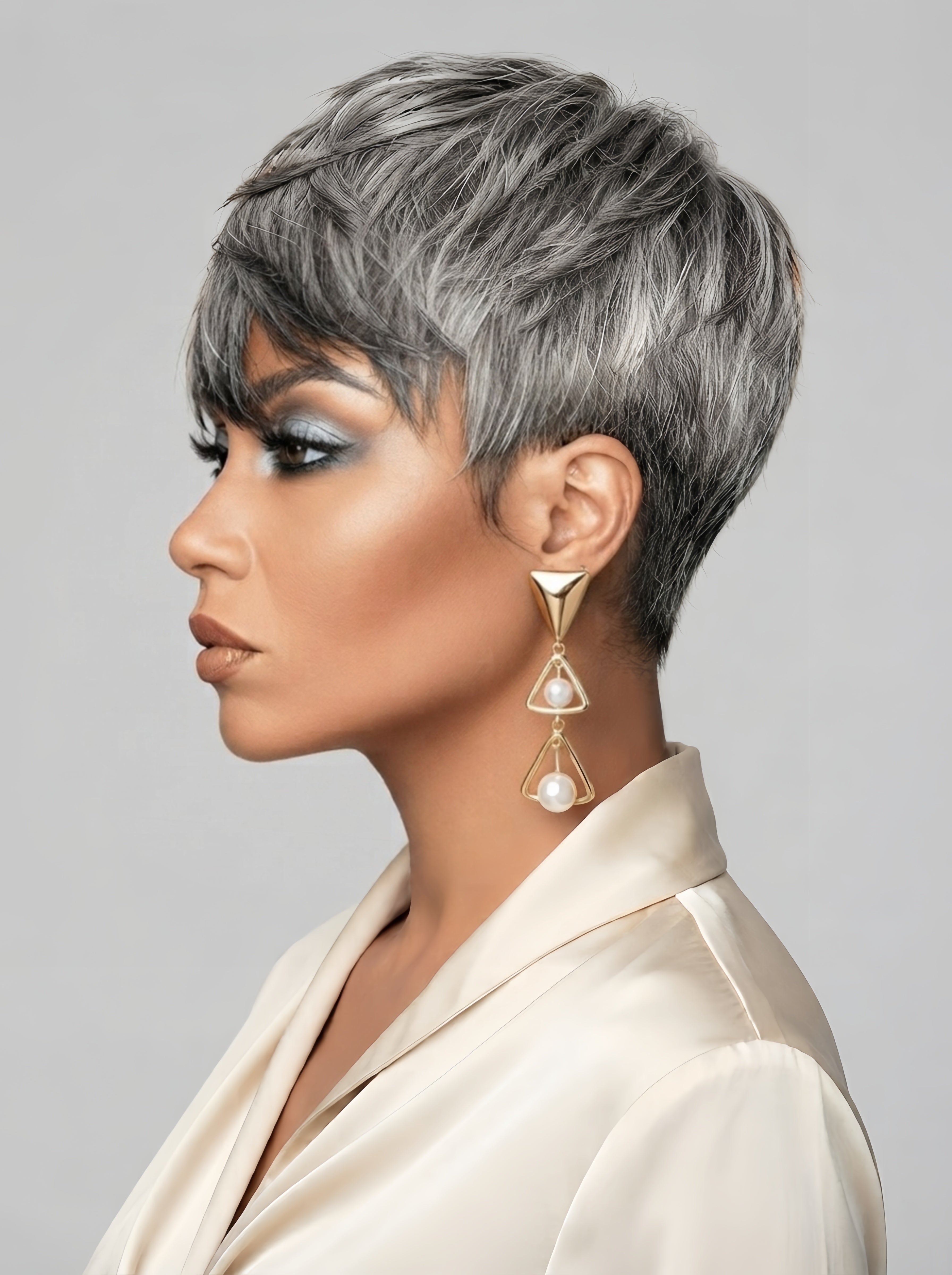 Side profile view of a model showcasing the tapered back and side layers of a grey pixie hair topper. The image highlights the volume at the crown and the smooth transition of the silver-grey strands. Her cool-toned blue eye makeup and gold triangular pearl earrings complement the modern, edgy vibe of the short hairstyle.