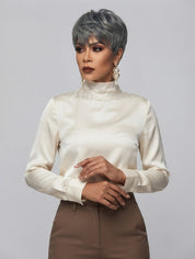 A model wearing a short, textured silver-gray wig, a cream satin mock-neck blouse, and brown trousers, posed against a solid light gray background.