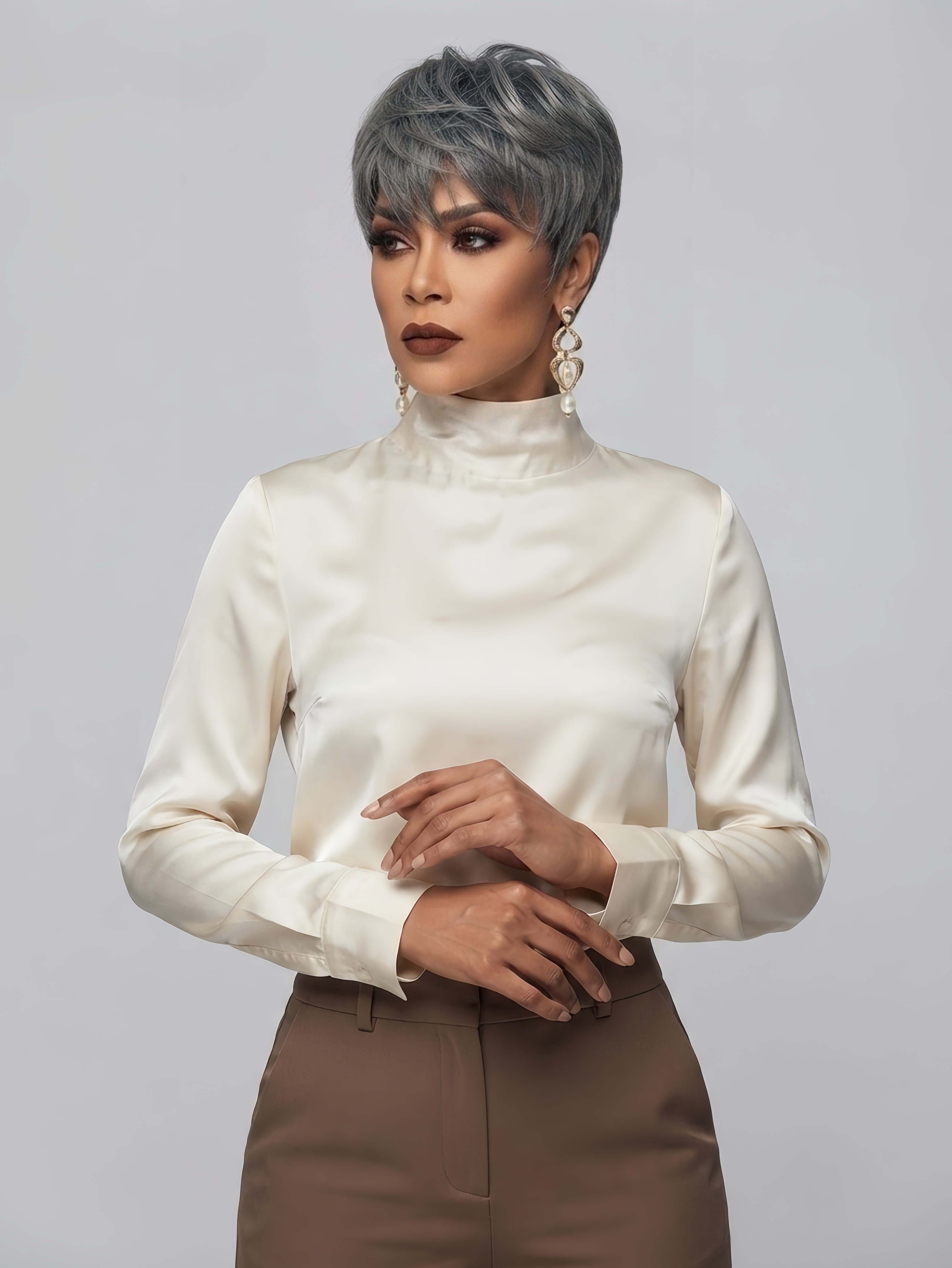A model wearing a short, textured silver-gray wig, a cream satin mock-neck blouse, and brown trousers, posed against a solid light gray background.