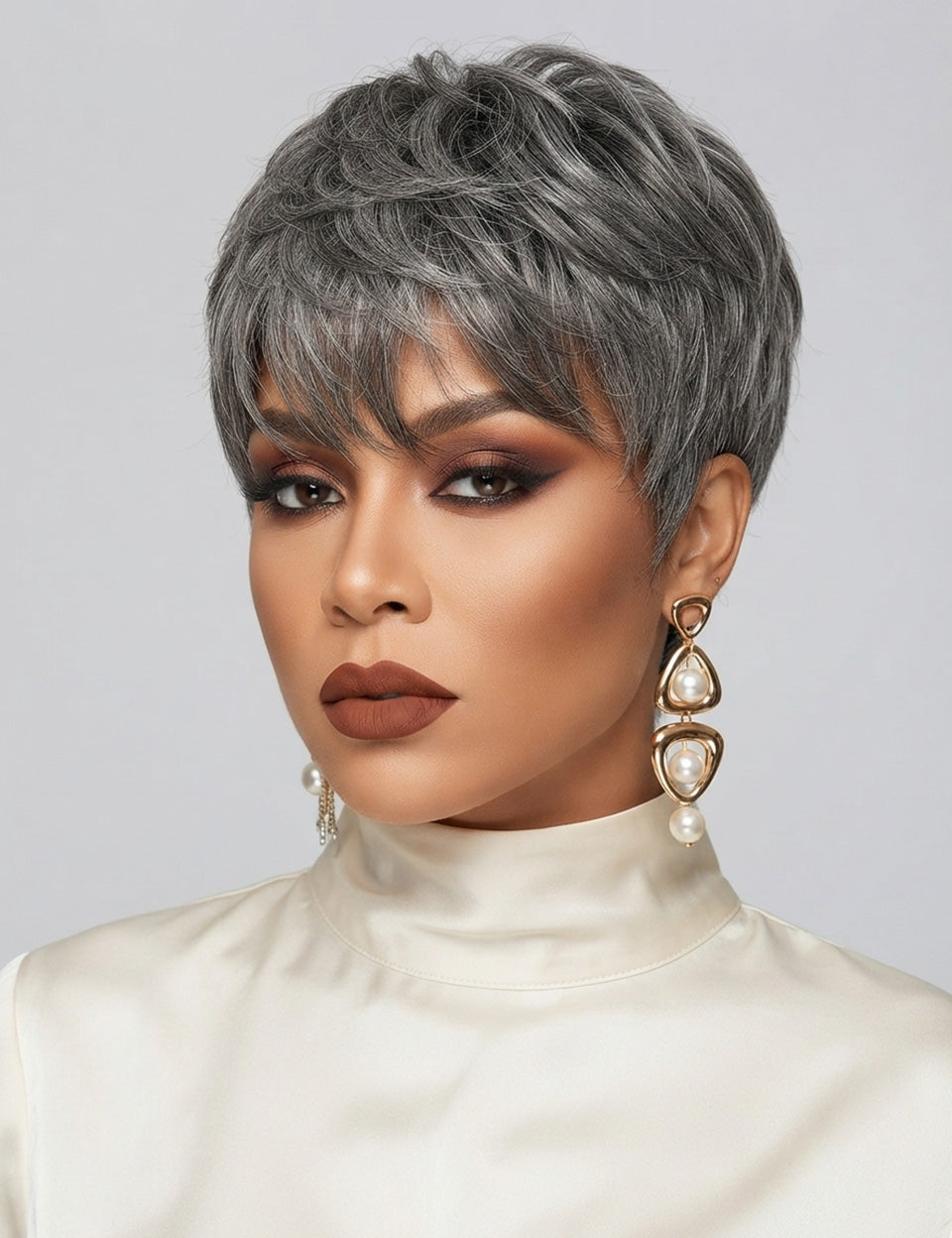 Three-quarter portrait of a woman in a chic salt and pepper pixie cut topper, featuring a soft side-swept fringe. The textured top adds height and dimension, while her bold terracotta eye makeup and matte brown lips enhance her features. Large gold hoop earrings with pearl accents complete the elegant and confident look.