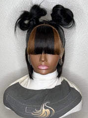 A black and brown two-tone wig displayed on a mannequin head with a white turtleneck, styled in a double braid top bun with bangs, set against a textured off-white wall.