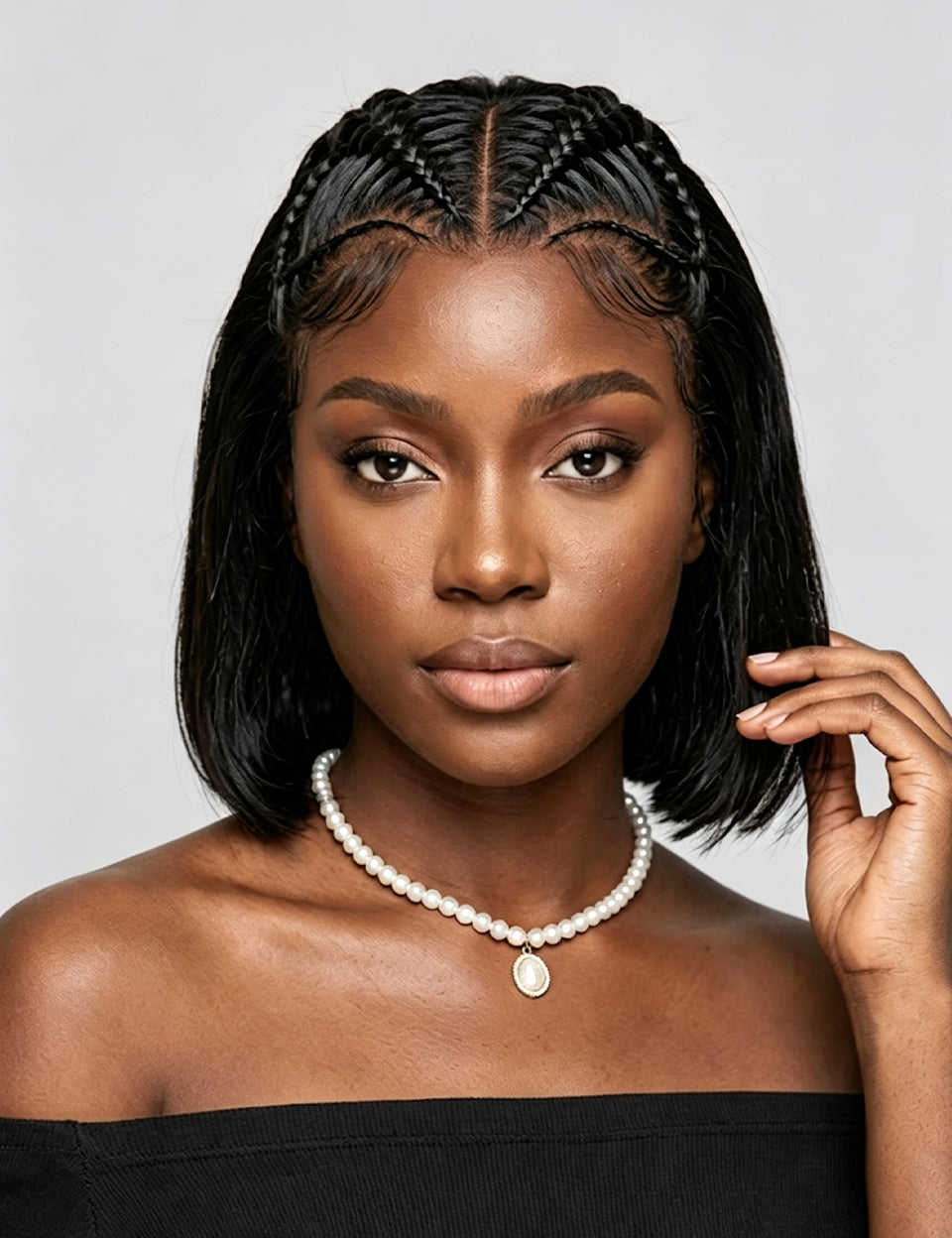 A woman with a short black bob haircut and braided hair details wears a pearl necklace with a white pendant, photographed against a light gray background.