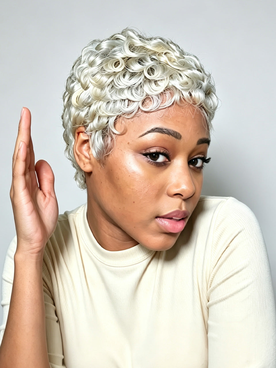 A close-up portrait of a woman wearing a short, tightly curled platinum blonde wig against a light gray studio background.