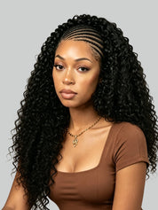 A woman with long, glossy, black curly hair styled in a deep wave pattern with a braided front part, wearing a brown scoop-neck top and a gold pendant necklace, set against a solid gray background.