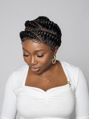 A woman with intricate black braided hair styled in a high updo, wearing a white long-sleeve top and gold jewelry, set against a solid light gray background.