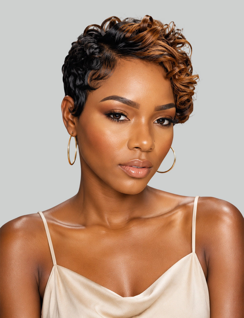 A woman with a short, curly, asymmetrical micro-lobed wig with dark brown and warm caramel highlights, wearing gold hoop earrings and a beige camisole against a solid gray background.
