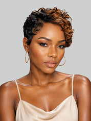 A woman with a short, curly, asymmetrical micro-lobed wig with dark brown and warm caramel highlights, wearing gold hoop earrings and a beige camisole against a solid gray background.